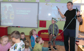 Grupa dzieci i mężczyzna. Mężczyzna prezentuje dzieciom sprzęt do wspinaczki górskiej 