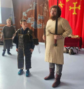 Mężczyzna i dwaj chłopcy. Wszyscy ubrani są w stroje historyczne. Trwa pokazowa lekcja dotycząca czasów pierwszych Piastów 