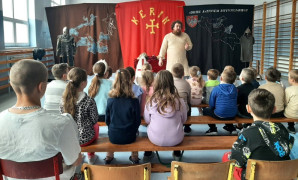 Dzieci siedzą na ławkach. Przed nimi stoi mężczyzna w historycznym stroju. Mężczyzna opowiada dzieciom o początkach państwa polskiego