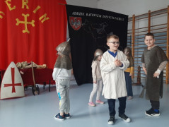 Dzieci stoją obok siebie. Trwa pokazowa lekcja dotycząca walk rycerskich. Dzieci ubrane są w historyczne stroje 