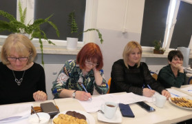 Cztery kobiety siedzą obok siebie przy stole. Kobiety trzymają w dłoniach długopisy