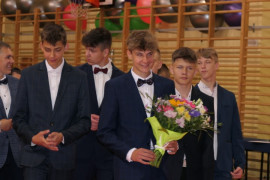 Grupa uczniów w garniturach. Jeden z nich trzyma bukiet kwiatów