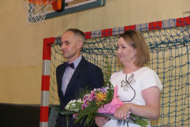 Kobieta - dyrektor szkoły - i mężczyzna  - nauczyciel  stoją obok siebie. Kobieta trzyma bukiet kwiatów