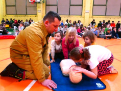 Dzieci uczestniczą w pokazowej akcji udzielania pierwszej pomocy. Asystuje im strażak 