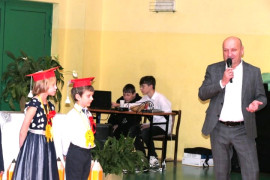 Mężczyzna trzyma w ręku mikrofon. Obok stoi  dwójka dzieci w czerwonych biretach 