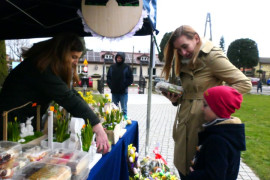 Stoisko wystawiennicze z wielkanocnymi ozdobami. Widoczne są osoby zainteresowane kupnem m.in. ciast i stroików