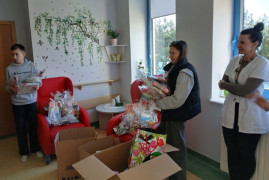 Kobieta, dziewczynka i chłopiec stoją obok siebie. Dziewczynka trzyma w dłoniach pudełko. Osoby znajdują się w pomieszczeniu 