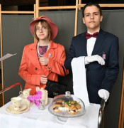 Dziewczyna w pomarańczowej marynarce oraz chłopak w czarny garniturze stoją obok siebie. Dziewczyna wcieliła się w rolę angielskiej królowej, natomiast chłopak wystąpił jako szarmancki lokaj. 