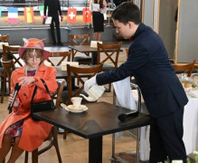 Scenka rodzajowa. Dziewczyna w pomarańczowej garsonce oraz w pomarańczowym kapeluszu siedzi przy stoliku. Chłopak w czarnym garniturze nalewa jej kawę 