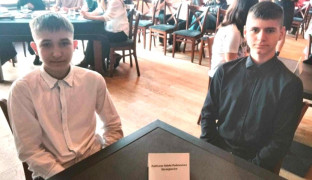 Dwóch chłopców siedzących przy stoliku. Chłopiec od lewej strony ubrany jest w białą koszulę, chłopiec siedzący z prawej strony ubrany jest w czarną koszulę 