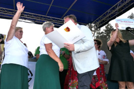 Dwie kobiety w zielonych długich spódnicach i mężczyzna w białej marynarce. Mężczyzna wręcza kobietom nagrodę 